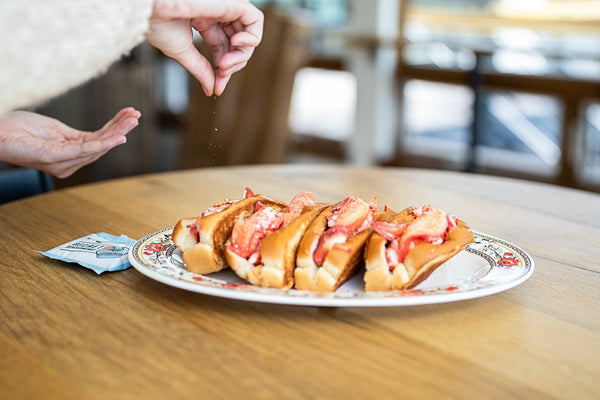 sprinkling seasoning over four lobster rolls on a plate