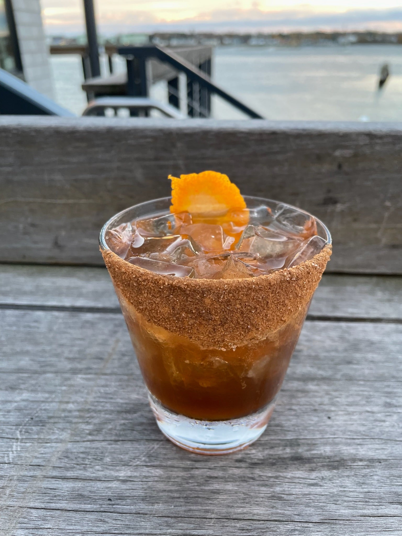a brown cocktail with a fruit wedge garnish on a deck overlooking the harbor