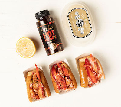 Half of a truffle lobster roll, half of a spicy honey butter lobster roll, and half of a traditional lobster roll on a white table next to the rolls' ingredients