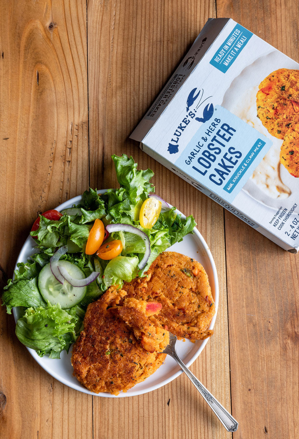a photo of lobster cakes on a plate with a side salad pictured below a package of lobster cakes